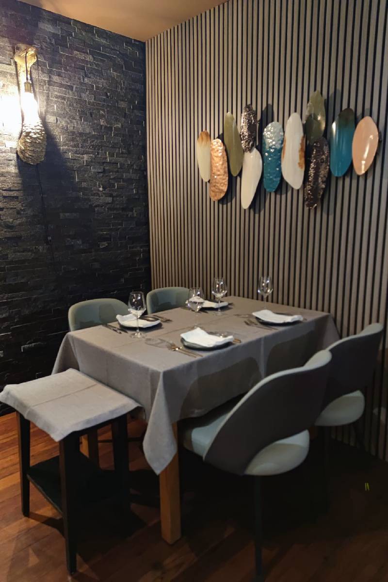 Mesa posta para quatro pessoas num canto acolhedor do restaurante Baco.come em Santa Maria da Feira, com parede de pedra escura e decoração moderna em peças metálicas na parede.