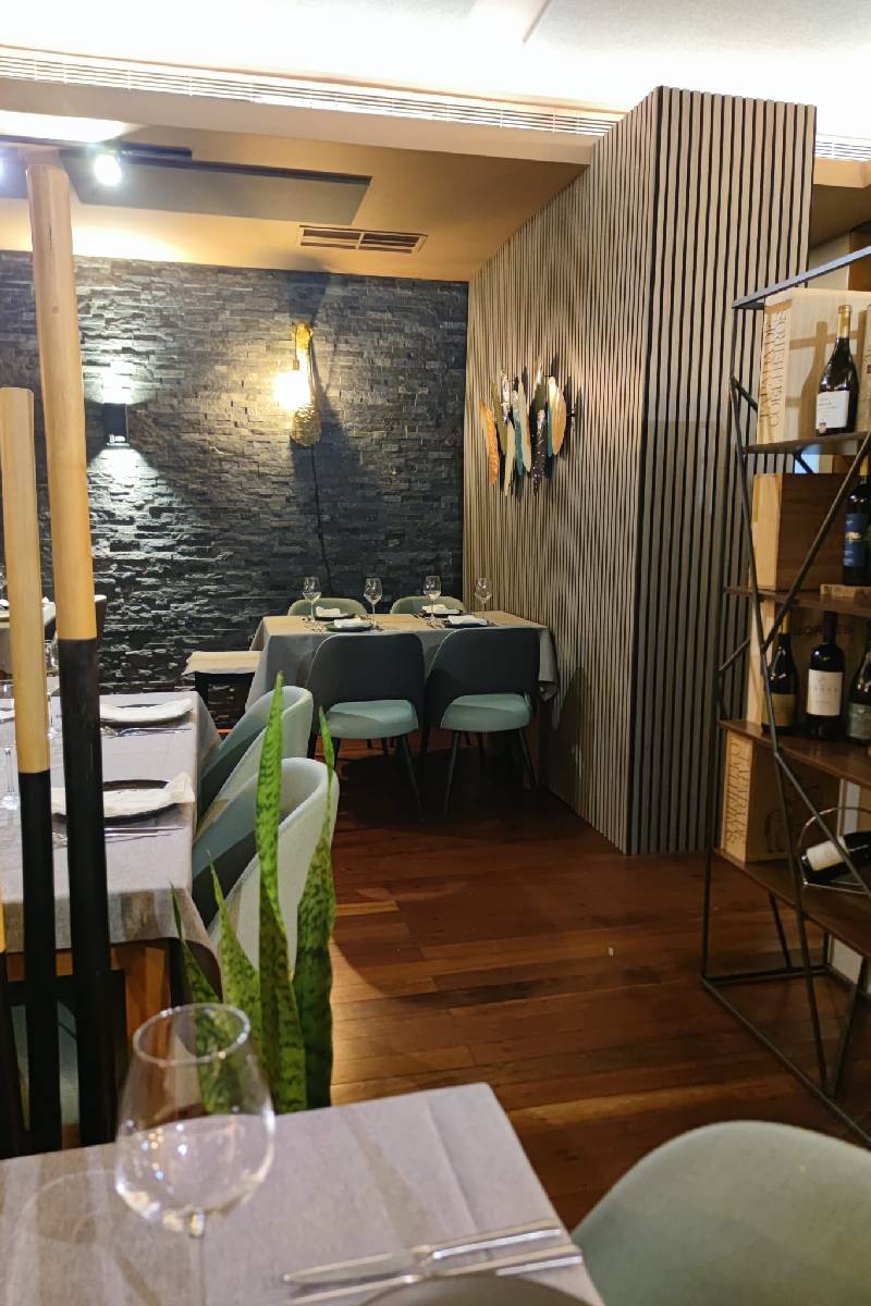 Vista interior do restaurante Baco.come em Santa Maria da Feira com mesas alinhadas, cadeiras em tons suaves e parede de pedra iluminada.